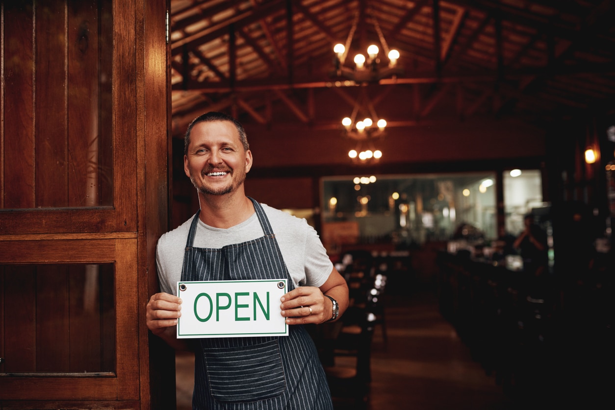 Portrait of a cheerful middle aged business owner holding up a sign saying "open" while standing under a doorway at a beer brewery during the day